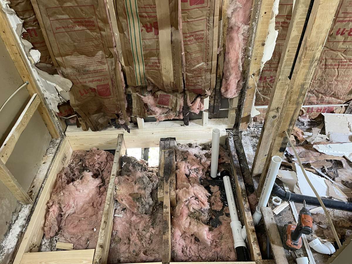 Bathroom stripped to the floor joists, showing rotted subfloor and framing with new drain and supply lines being run